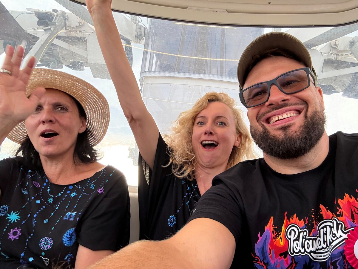 Three people - two women and a man - are sitting in the gondola of a Ferris wheel. The man is taking a selfie, the women have their hands raised. In the background you can see the construction of the mill.